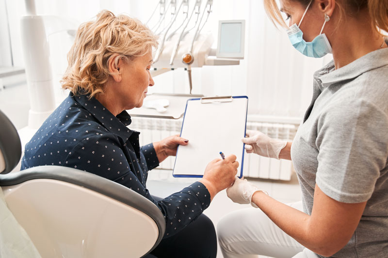 Side view of the female patient with a pen in her hand signing the medical application form of her new dentist sitting in front of her.