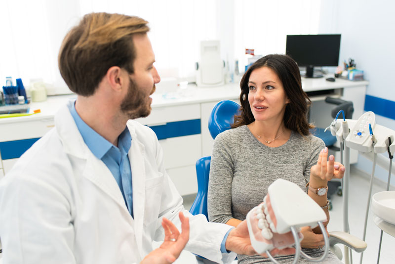 The image depicts a dental professional showing a dental model to a patient. The patient, a woman, is attentively listening and engaging in conversation with the dentist. They are in a well-equipped dental office, suggesting a consultation or discussion about dental treatment options.