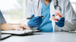 A dentist wearing blue gloves explains a dental procedure using a tooth model, while a patient takes notes on a clipboard, illustrating a consultation session.