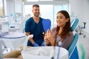 Happy woman looking at her teeth in a hand held mirror at dentist's office, male dentist interacting with the female patient.