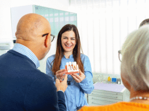 Female dentist holding anatomical model of teeth and explaining dental implant procedure to senior patients in clinic.